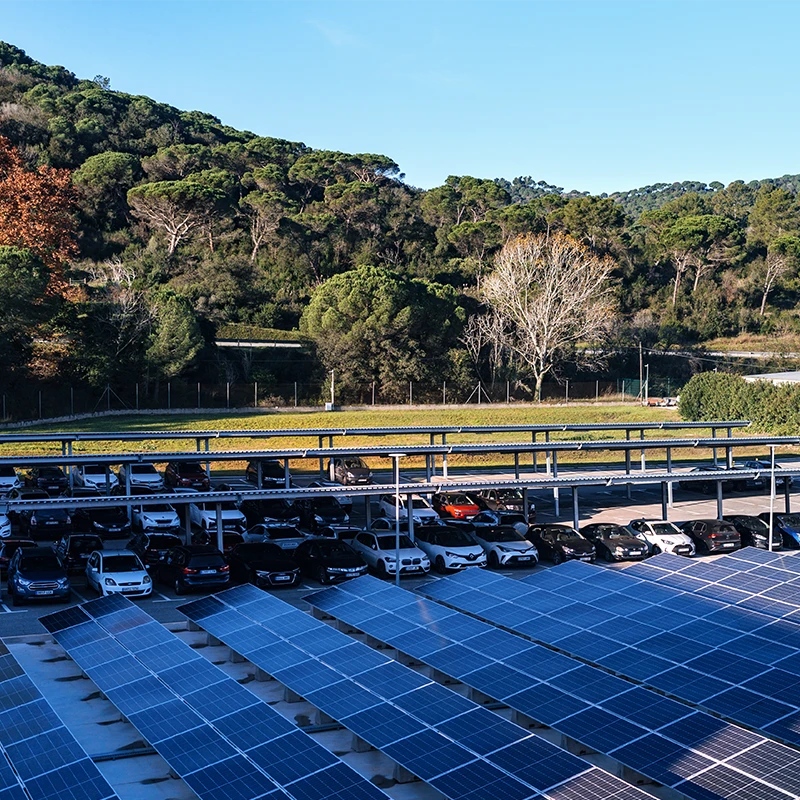 In the image, there is a large parking lot filled with cars, with a solar panel array located nearby. The parking lot seems to be outdoors and surrounded by nature, possibly near a park or a forest. (photo)